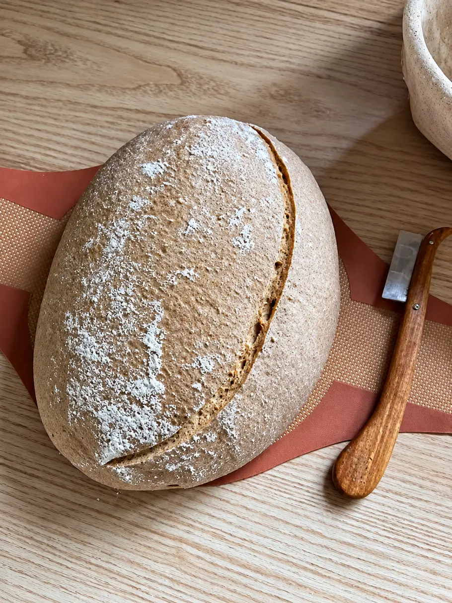 am nächsten Tag Brot einschneiden Ein bemehlter, ovaler Brotlaib mit langem Einschnitt liegt auf einer Backmatte, daneben eine Klinge mit Holzgriff.