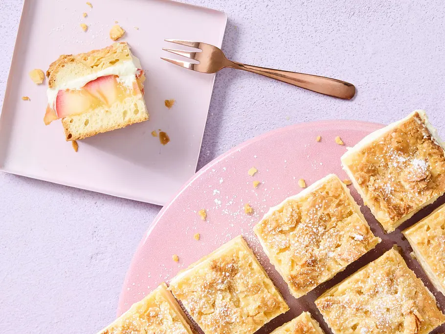 Rechteckige Stücke Apfel-Bienenstich mit goldbrauner Mandelkruste und Puderzucker, angerichtet auf einer rosa Platte, ein Stück auf einem kleinen Teller.