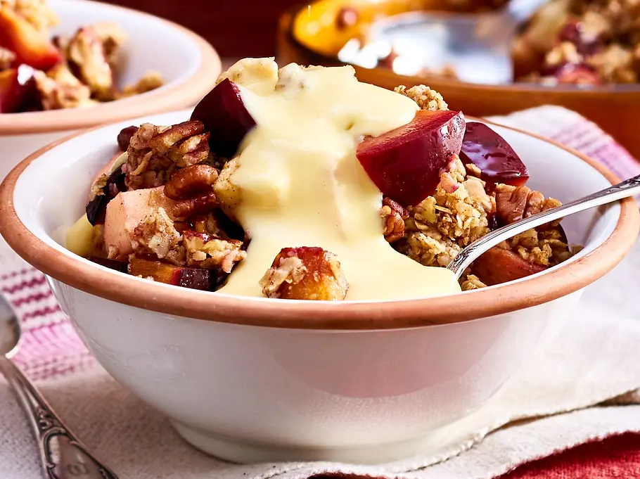 Warmer Apfel-Pflaumen-Crumble mit knusprigen Streuseln und Pekannüssen in einer weißen Schale, großzügig mit Vanillesoße übergossen. Im Hintergrund stehen weitere Portionen sowie eine Form mit dem restlichen Crumble.