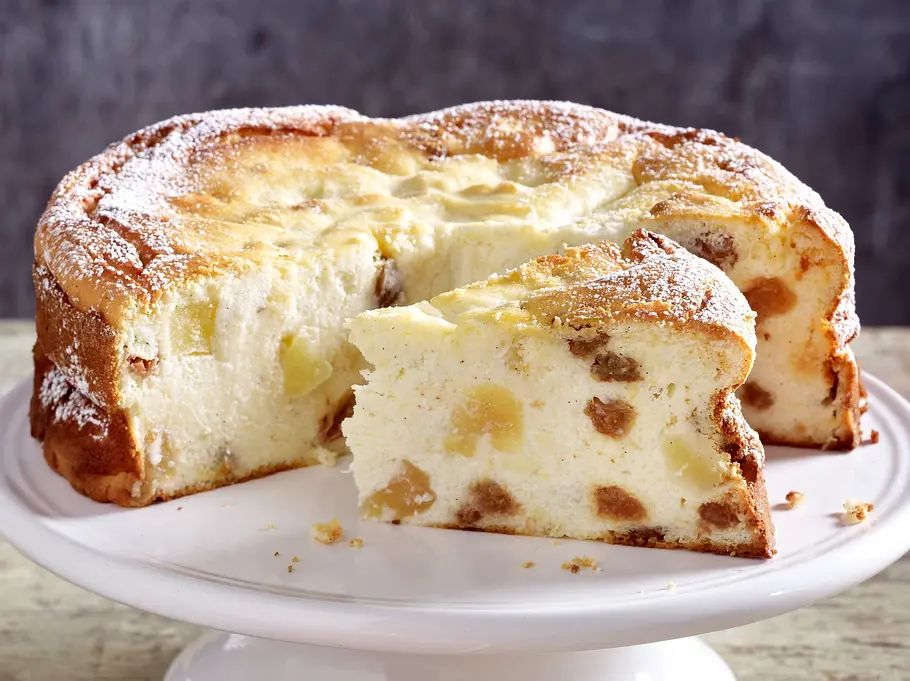 Apfel-Quarkkuchen Rezept Frank Rosin Der Apfel-Quarkkuchen ist auf einer weißen Tortenplatte serviert und angeschnitten. Man kann Rosinen und Apfelstückchen erkennen.
