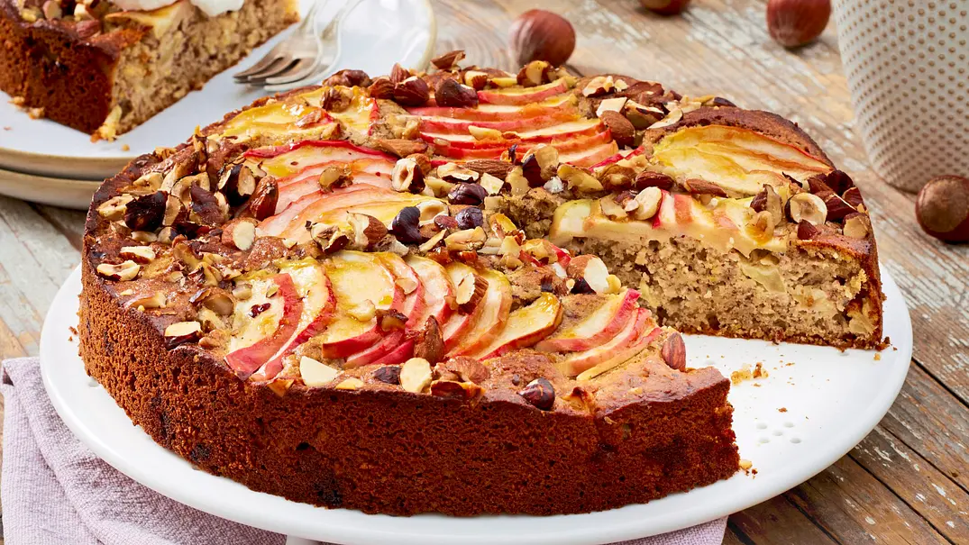 Runder Apfel-Vollkorn-Nusskuchen aus der Springform, aus dem ein großes Stück herausgeschnitten wurde auf einem weißen Kuchenteller - Foto: LECKER @ Bauer Media Group Runder Apfel-Vollkorn-Nusskuchen aus der Springform, aus dem ein großes Stück herausgeschnitten wurde auf einem weißen Kuchenteller - Foto: LECKER @ Bauer Media Group