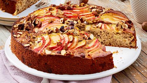 Runder Apfel-Vollkorn-Nusskuchen aus der Springform, aus dem ein großes Stück herausgeschnitten wurde auf einem weißen Kuchenteller - Foto: LECKER @ Bauer Media Group