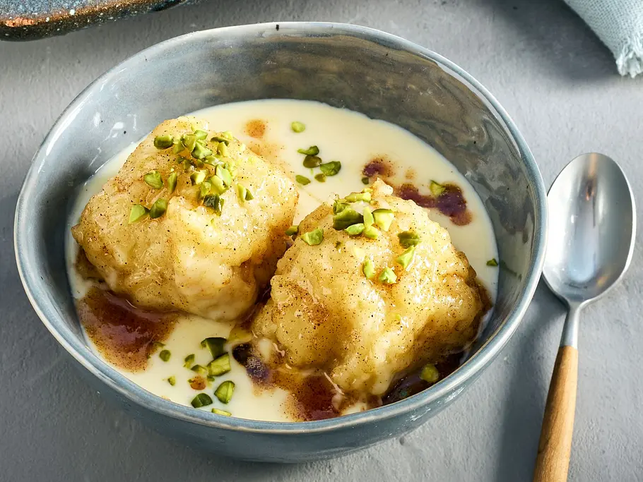 Apfelknödel sind in einer Schüssel mit Vanillesoße und gehackten Pistazienkernen angerichtet.
