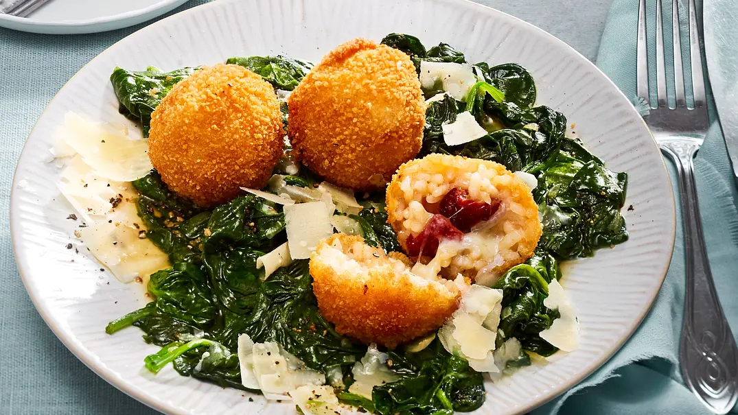 Drei knusprig gebackene Arancini auf einem Bett aus frischem Blattspinat und Parmesanraspeln, einer der Arancini ist aufgeschnitten und zeigt eine Reisfüllung mit Tomate und Mozzarella, serviert auf einem weißen Teller. - Foto: LECKER @ Bauer Media Group Drei knusprig gebackene Arancini auf einem Bett aus frischem Blattspinat und Parmesanraspeln, einer der Arancini ist aufgeschnitten und zeigt eine Reisfüllung mit Tomate und Mozzarella, serviert auf einem weißen Teller. - Foto: LECKER @ Bauer Media Group