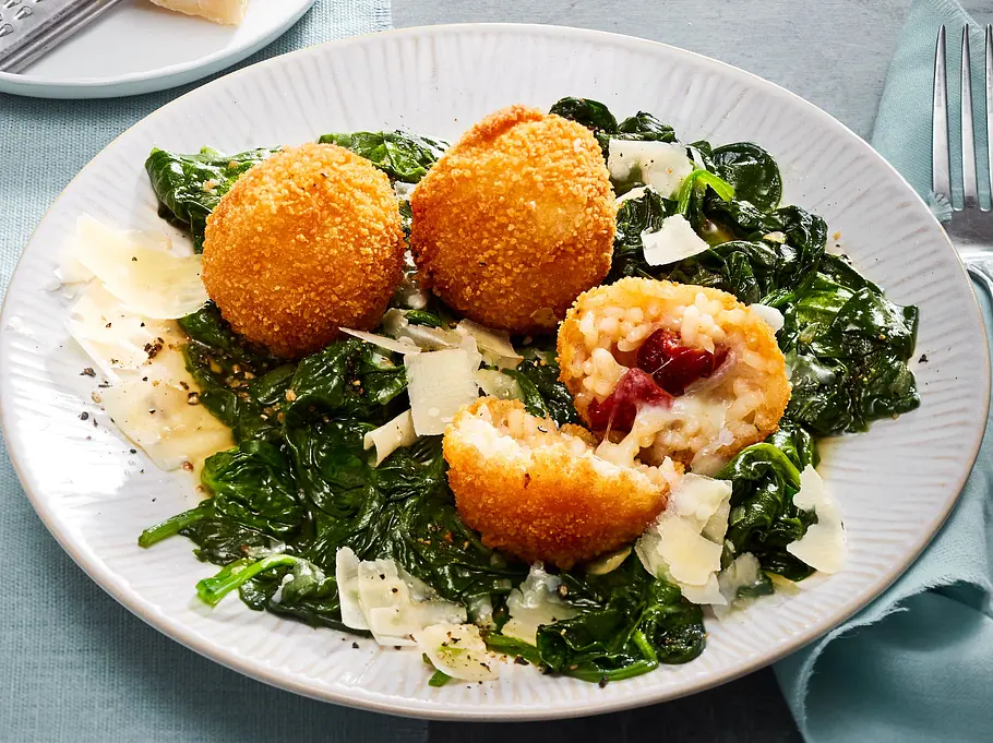 Drei knusprig gebackene Arancini auf einem Bett aus frischem Blattspinat und Parmesanraspeln, einer der Arancini ist aufgeschnitten und zeigt eine Reisfüllung mit Tomate und Mozzarella, serviert auf einem weißen Teller.