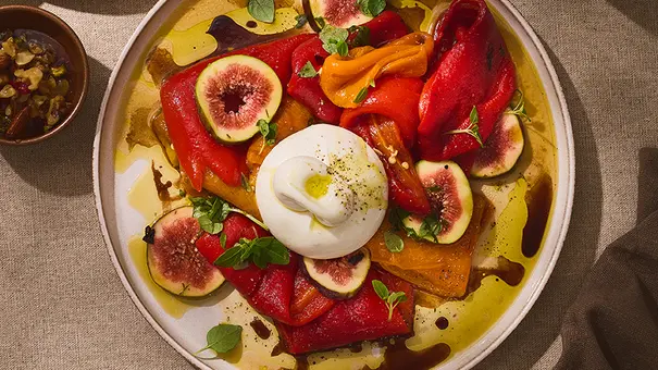 Cremige Burrata auf einem runden, hellen Teller, umgeben von bunten eingelegten Paprikaschoten, frischen Feigenscheiben und Kräutern, beträufelt mit Olivenöl und Balsamico. - Foto: ITA Assolatte