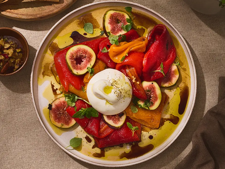 Burrata mit eingelegten Paprikaschoten Cremige Burrata auf einem runden, hellen Teller, umgeben von bunten eingelegten Paprikaschoten, frischen Feigenscheiben und Kräutern, beträufelt mit Olivenöl und Balsamico.