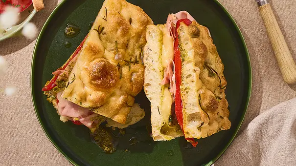 Auf einem dunkelgrünen Teller liegt ein aufgeschnittenes Focaccia-Sandwich, gefüllt mit Mortadella, geschmolzenem Provolone, eingelegter Paprika und etwas grünem Pesto. - Foto: ITA Assolatte