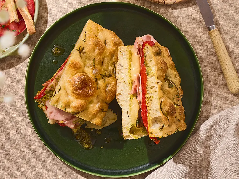 Focaccia-Sandwich mit Paprika, Mortadella & Provolone Auf einem dunkelgrünen Teller liegt ein aufgeschnittenes Focaccia-Sandwich, gefüllt mit Mortadella, geschmolzenem Provolone, eingelegter Paprika und etwas grünem Pesto.