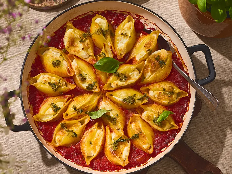 Gefüllte Pasta mit Zitronen-Ricotta & Pesto Gefüllte Pasta-Muscheln in einer runden Auflaufform, gefüllt mit cremiger Zitronen-Ricotta-Mischung, serviert auf einer Schicht Tomatensauce und garniert mit grünem Pesto und frischem Basilikum.