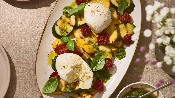 Grillgemüse wie Zucchini und gelbe Paprika, angerichtet auf einer ovalen Platte, getoppt mit zwei Kugeln Mozzarella di Bufala, frischem Basilikum und grüner Kräutersoße. - Foto: ITA Assolatte