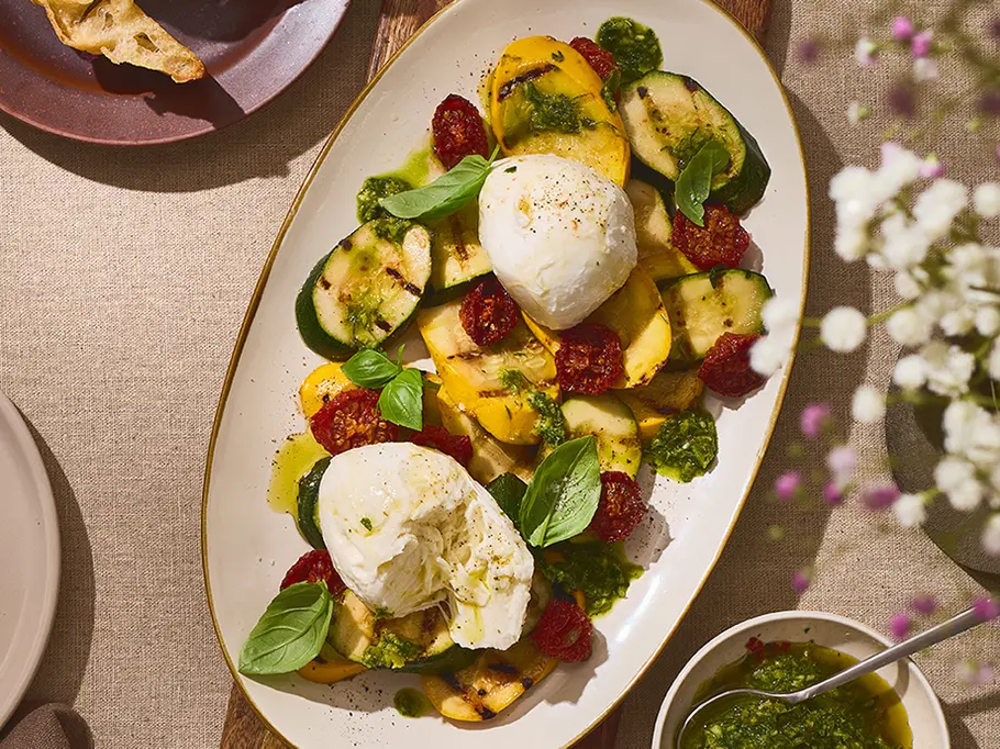 Gegrilltes Gemüse mit Mozzarella di Bufala Grillgemüse wie Zucchini und gelbe Paprika, angerichtet auf einer ovalen Platte, getoppt mit zwei Kugeln Mozzarella di Bufala, frischem Basilikum und grüner Kräutersoße.