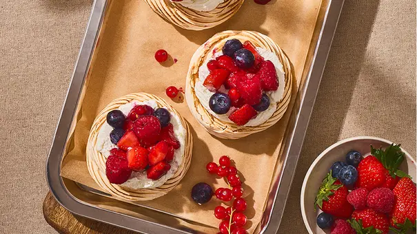 Drei kleine Pavlovas mit cremiger Mascarpone-Füllung und bunt gemischten Beeren, angerichtet auf Backpapier in einer eckigen Blechform. - Foto: ITA Assolatte