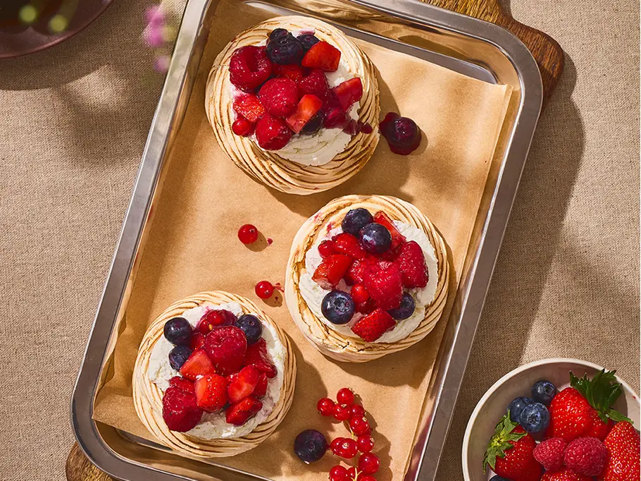 Kleine Pavlova mit Mascarpone-Creme und Beeren Drei kleine Pavlovas mit cremiger Mascarpone-Füllung und bunt gemischten Beeren, angerichtet auf Backpapier in einer eckigen Blechform.