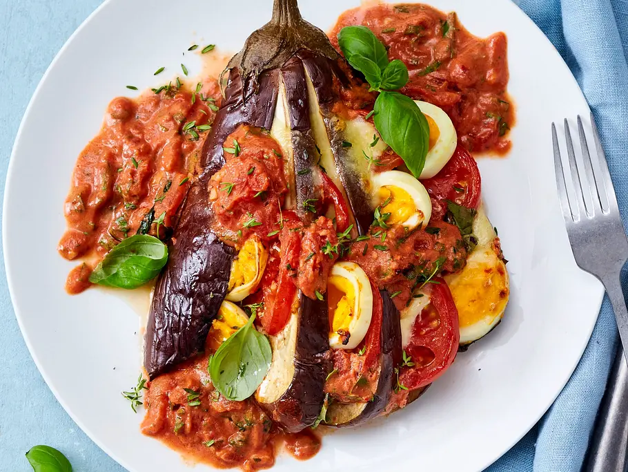 Gebackene Aubergine, fächerförmig eingeschnitten und mit Tomaten- und Käsescheiben gefüllt, liegt in kräftiger Tomatensoße auf einem weißen Teller. Frischer Basilikum und eine blaue Serviette runden das Gericht ab.