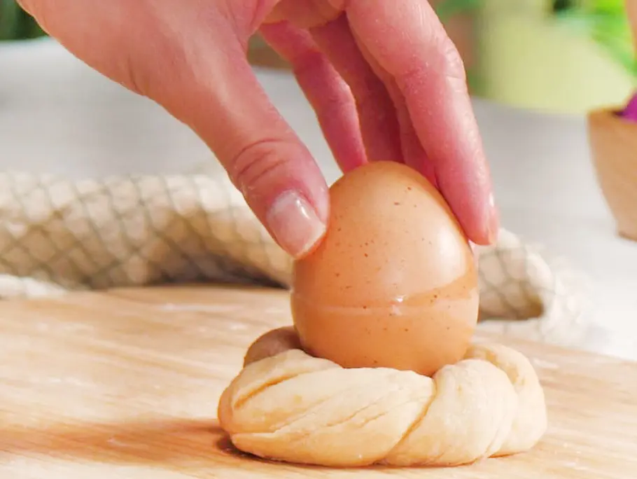 Ungebackener, geflochtener Hefeteigkranz, in dessen Mitte ein braunes Ei gesetzt wird; Hand platziert das Ei auf einem Holzbrett.