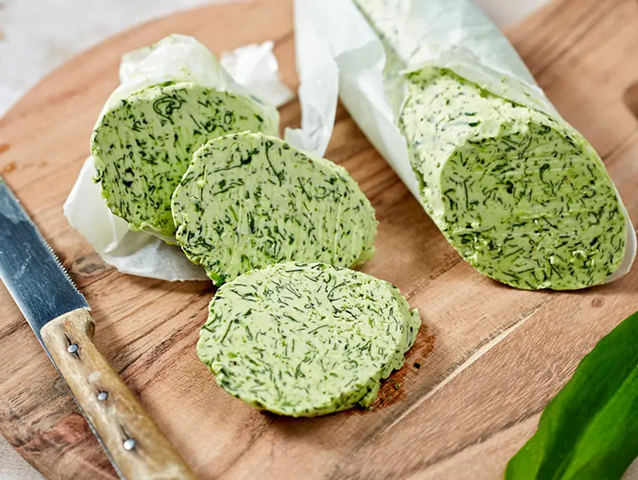 Bärlauchbutter-Rezept Eine Rolle Bärlauchbutter im Backpapier liegt auf einem Holzbrett, daneben drei Scheiben, ein Messer und ein Bärlauchblatt.