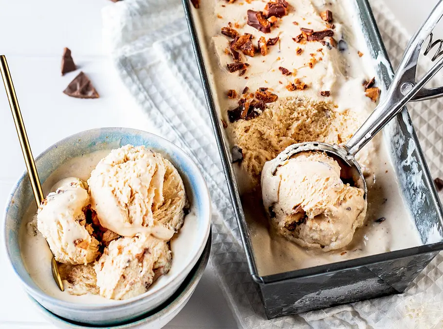 Baileys-Eis-Rezept Eine Schale mit Löffel voll mit heller Eiscreme, daneben eine Kastenform mit mehr Eis getoppt mit Stückchen und einem Portionierer, der eine Eiskugel formt.