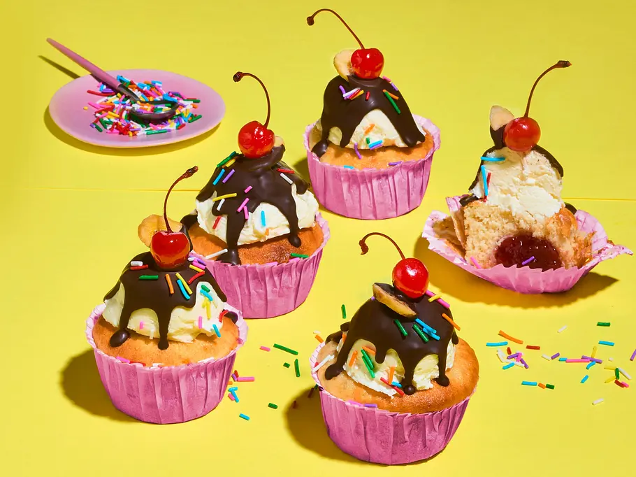 Banana Split Muffins Fünf Cupcakes in rosa Förmchen mit Eis, Schokoguss, bunten Streuseln und Kirschen stehen auf gelbem Untergrund.