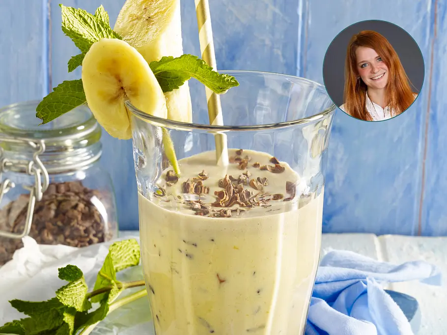 Bananen-Mandel-Smoothie getoppt mit Kakaonibs in einem hohen Glas mit einer Deko aus einer Bananenscheibe, einem länglichen Stück Banane und frischer Minze. Mit im Bild Redakteurin Mareen.