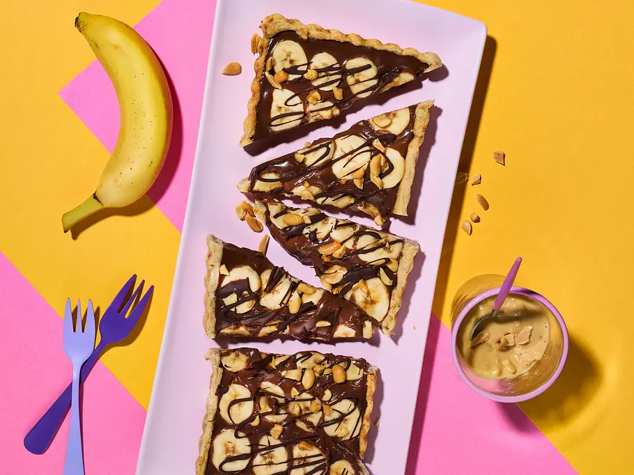 Bananen-Schoko-Tarte mit Erdnuss-Crunch Schoko-Bananen-Tarte liegt auf einer länglichen Platte, daneben Banane, Gabeln und ein Glas Creme auf gelb-pinkem Untergrund.