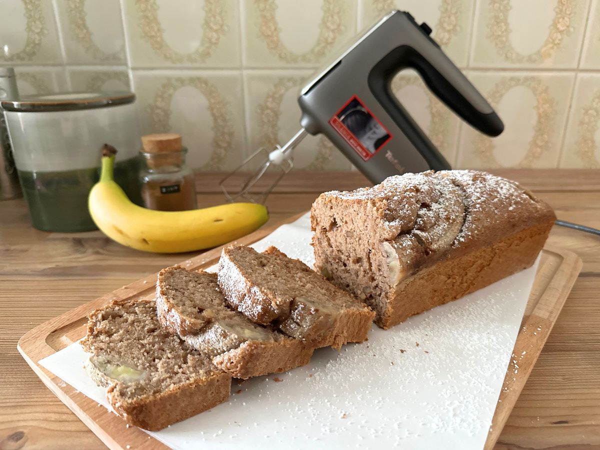 Ein angeschnittenes Bananenbrot steht auf einem Küchenbrett vor dem Tefal-Handmixer