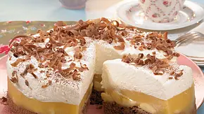 Bananentorte mit lockerer Sahneschicht, einer Schicht Puddingcreme und frischen Bananen auf einem dunklen Biskuitboden, garniert mit Schokoraspeln, serviert auf einem rosa Teller. - Foto: LECKER @ Bauer Media Group