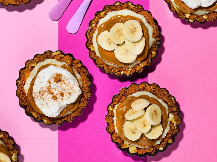 Banoffee-Tartelettes Sieben Törtchen mit Bananenscheiben oder Sahne und zwei rosa Gabeln liegen auf einem rosa-pink gemusterten Untergrund.