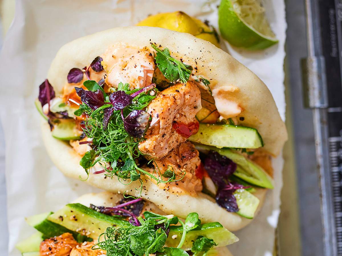 Saftige Bao Buns gefüllt mit zartem Lachs, frischen Gurkenstreifen, Kräutern, fein abgeschmeckt mit cremiger Sauce und knackigen Microgreens, serviert auf hellem Backpapier mit Limetten im Hintergrund.