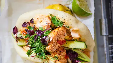 Saftige Bao Buns gefüllt mit zartem Lachs, frischen Gurkenstreifen, Kräutern, fein abgeschmeckt mit cremiger Sauce und knackigen Microgreens, serviert auf hellem Backpapier mit Limetten im Hintergrund. - Foto: LECKER @ Bauer Media Group