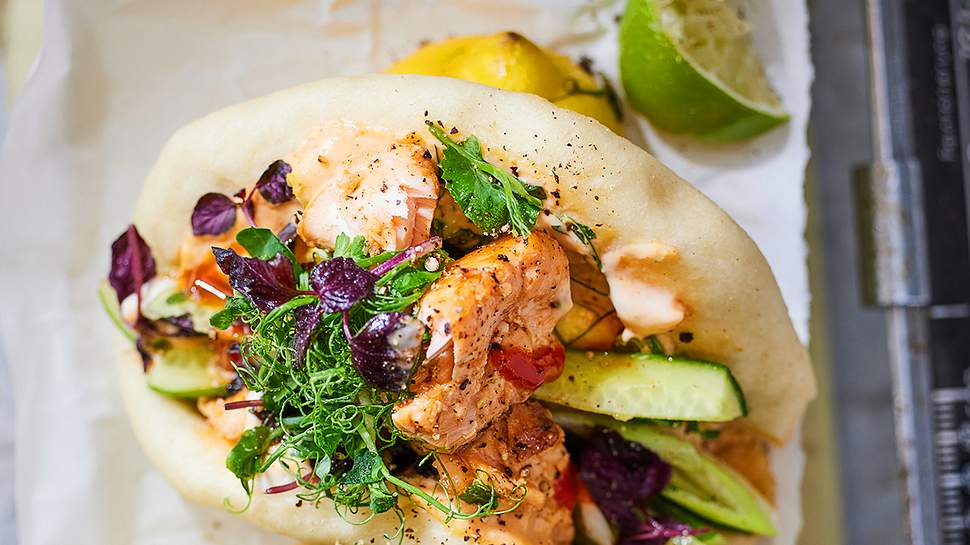 Saftige Bao Buns gefüllt mit zartem Lachs, frischen Gurkenstreifen, Kräutern, fein abgeschmeckt mit cremiger Sauce und knackigen Microgreens, serviert auf hellem Backpapier mit Limetten im Hintergrund. - Foto: LECKER @ Bauer Media Group