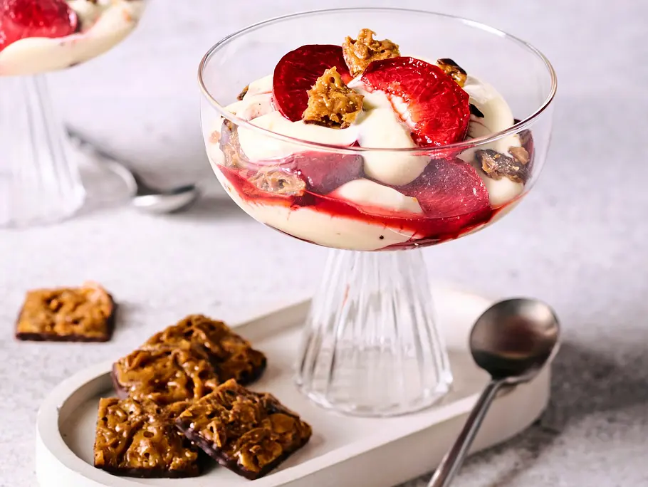 Bienenstich-Dessert mit cremiger Schicht, frischen Pflaumenstücken und knusprigen Mandelkeksen in eleganten Glasschalen, serviert auf hellem Untersetzer mit Löffel und zusätzlichem Gebäck.