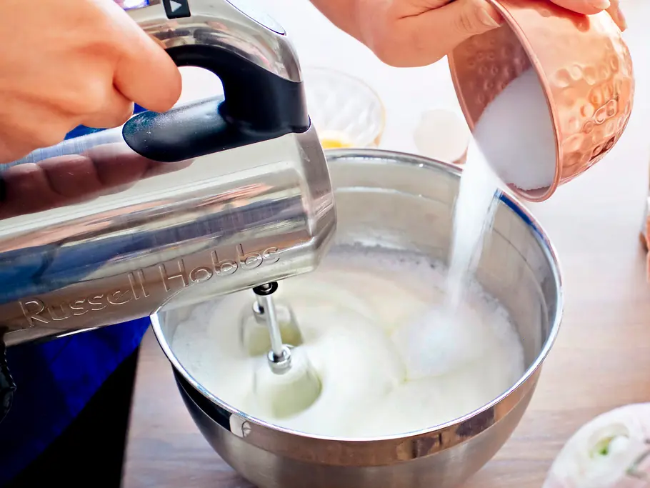 Eischnee wird in einer Metallschüssel mit einem elektrischen Handmixer geschlagen, während Zucker aus einer kupferfarbenen Schale langsam eingerieselt wird; auf der Holzplatte stehen weitere Zutaten und Küchenutensilien.