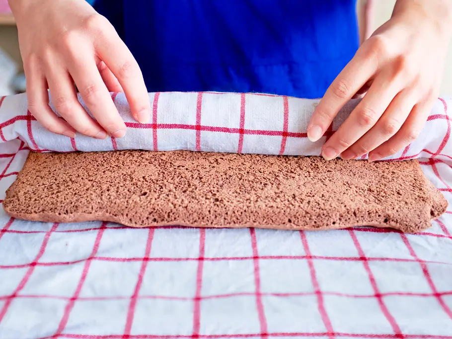 Frisch gebackener Schokoladen-Biskuitteig wird auf einem rot-weiß karierten Geschirrtuch von Hand eingerollt.