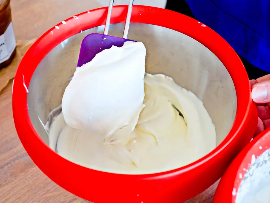 Locker aufgeschlagene Schmand-Sahne-Creme wird mit einem Spatel in einer silbernen Rührschüssel mit rotem Rand verrührt, auf einer Holzarbeitsfläche.