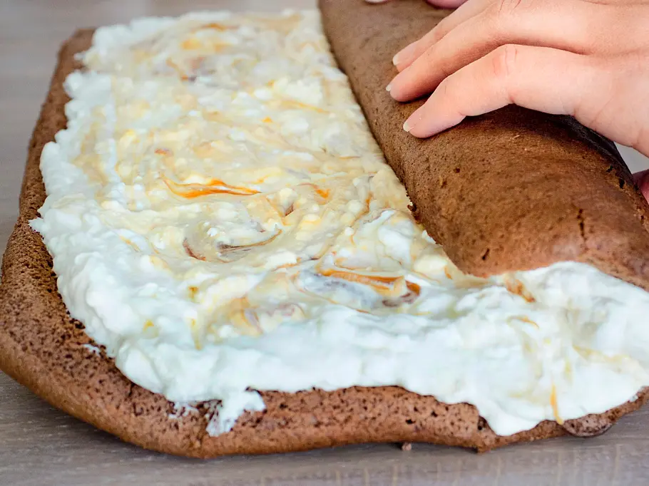 Schokoladen-Biskuitteig wird mit einer hellen Sahne-Karamell-Creme bestrichen und von Hand auf einer Holzplatte aufgerollt.