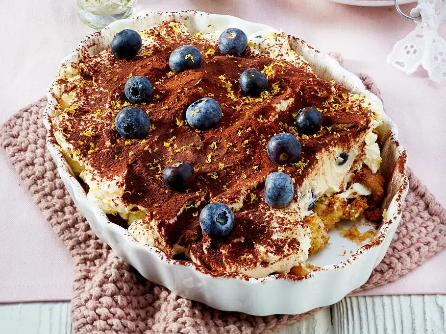 Blaubeer-Buttermilch-Tiramisu Cremiges Blaubeer-Buttermilch-Tiramisu garniert mit frischen Blaubeeren und Kakaopulver in einer weißen Auflaufform.