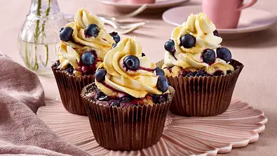 Drei saftige Blaubeer-Cupcakes mit cremigem Orangen-Frosting und frischen Blaubeeren als Topping, angerichtet auf einer pastellrosa Schale. - Foto: LECKER @ Bauer Media Group