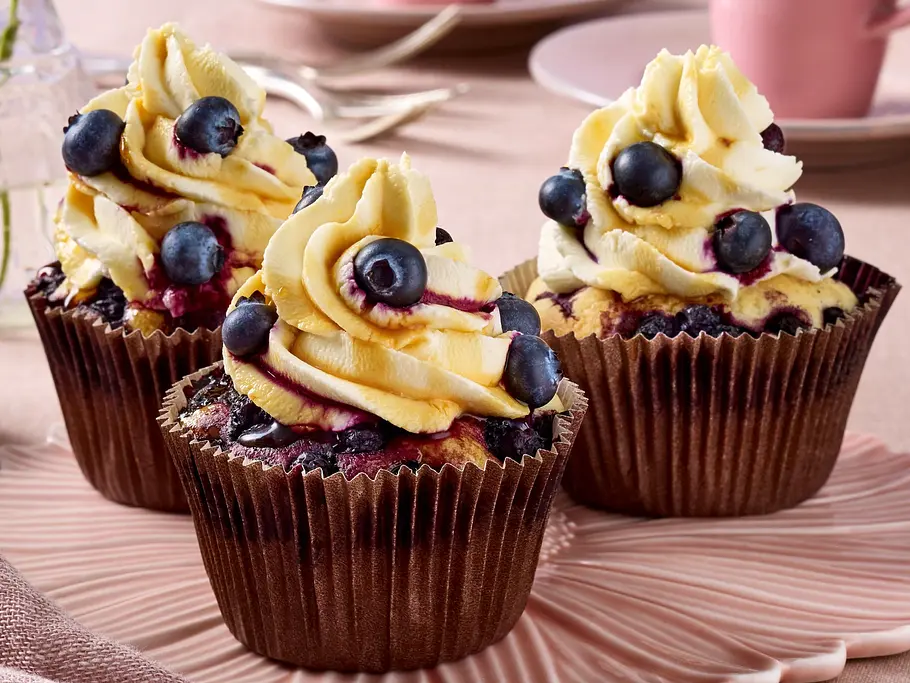 Drei saftige Blaubeer-Cupcakes mit cremigem Orangen-Frosting und frischen Blaubeeren als Topping, angerichtet auf einer pastellrosa Schale.