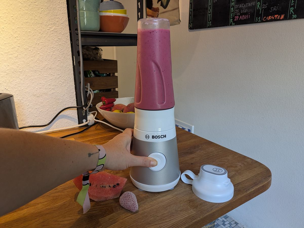 Bosch Smoothie-Maker VitaPower Serie 2 Knopf Der Bosch VitaPower Serie 2 Smoothie-Maker wird mit einer Hand bedient, daneben liegen Deckel, Melonenscheibe und gefrorene Erdbeere.