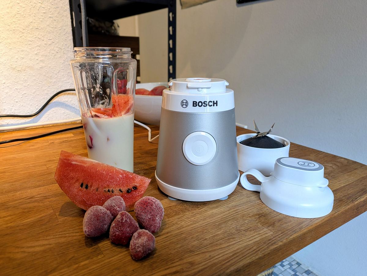 Bosch Smoothie-Maker VitaPower Serie 2 Der Bosch VitaPower Serie 2 Smoothie-Maker mit Mixbecher voller Milch, Melone und Früchten steht auf Holzplatte, daneben Deckel und Zubehör.