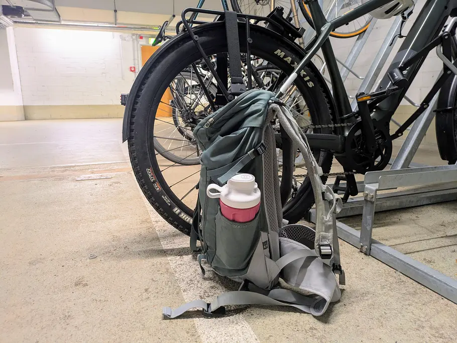 Ein Rucksack steht in einem Fahrradkeller, in der Seitentasche steckt ein Smoothiebecher mit pinkem Getränk.