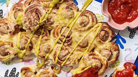 Herzhaftes Zupfbrot aus Teigschnecken mit Bratwurstfüllung und geschmolzenem Käse, auf einem Tablett mit bunter Serviette angerichtet, dazu Senf und Tomatensauce in einer Schale. - Foto: Image Professionals