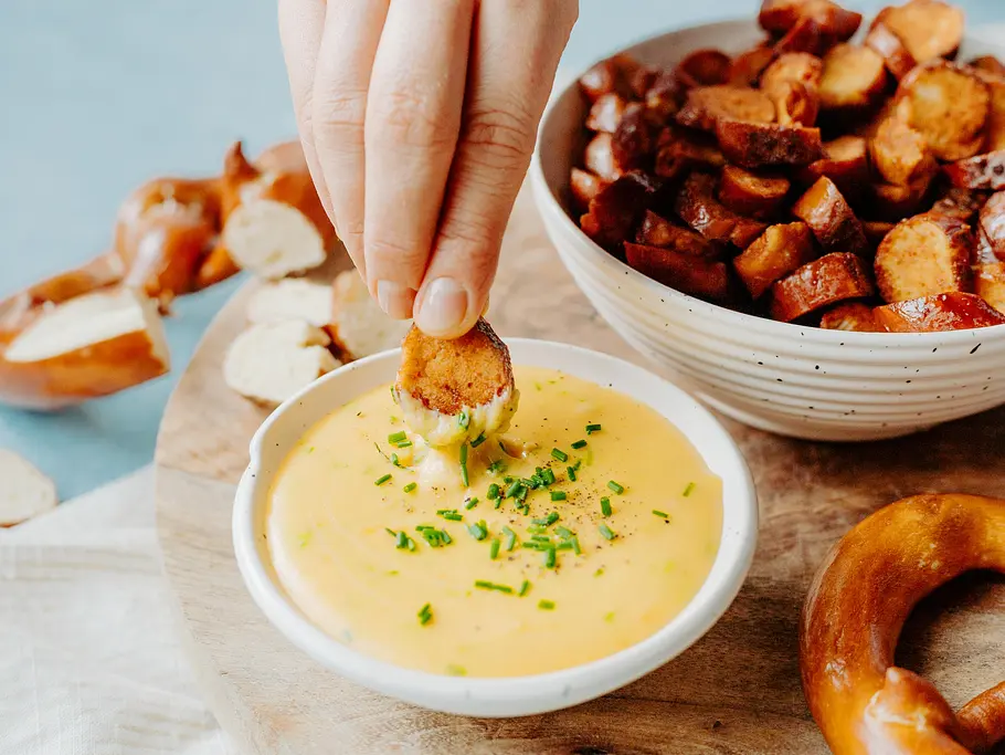 Eine Hand taucht ein geröstetes Laugengebäckstück in gelben Käse-Dip mit Schnittlauch. Daneben steht eine Schüssel mit weiteren Stücken.
