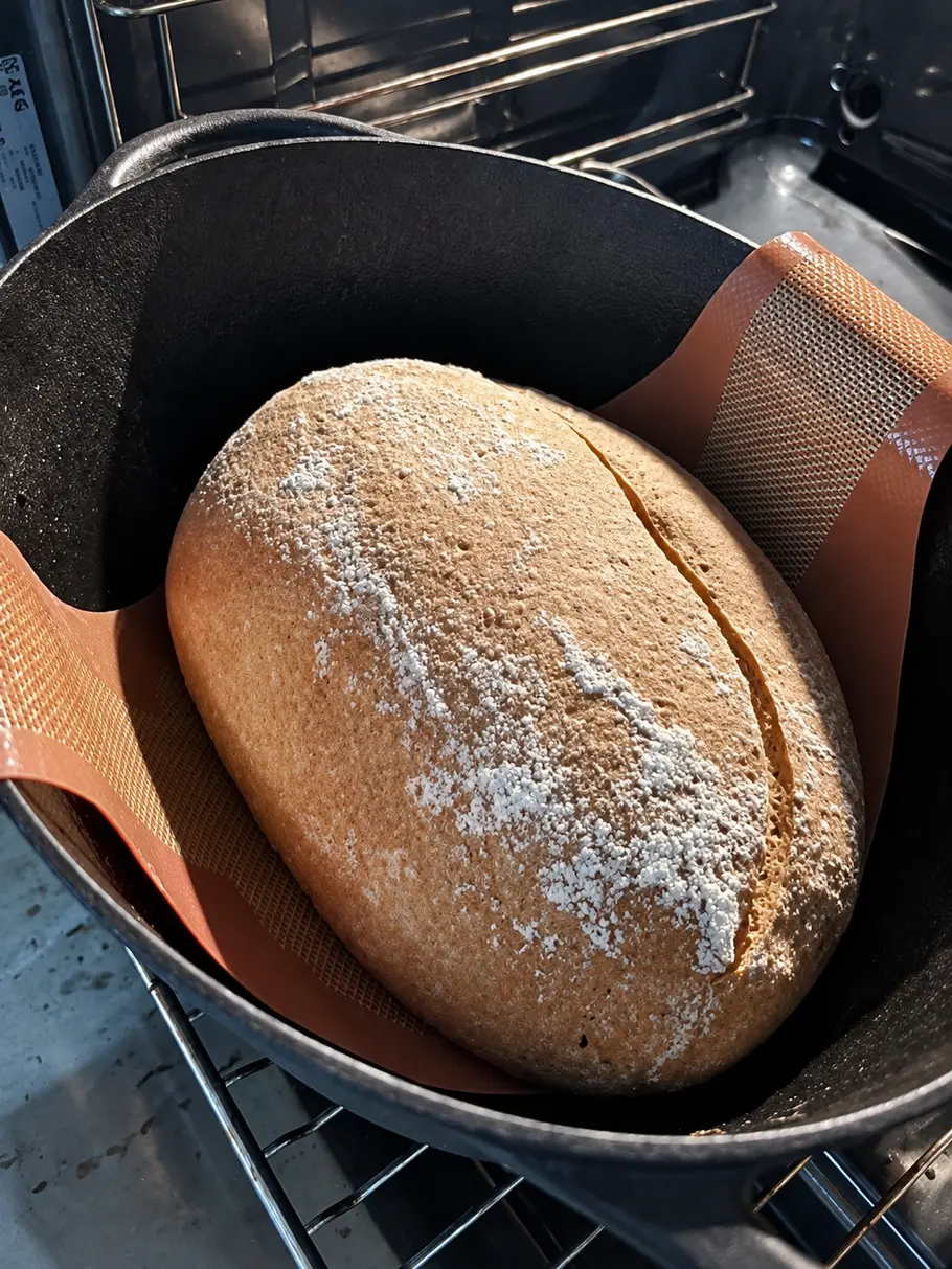 Brotlaib backen Ein bemehltes, längs eingeschnittenes Brot liegt auf einer rotbraunen Backmatte in einem schwarzen Topf im Backofen.