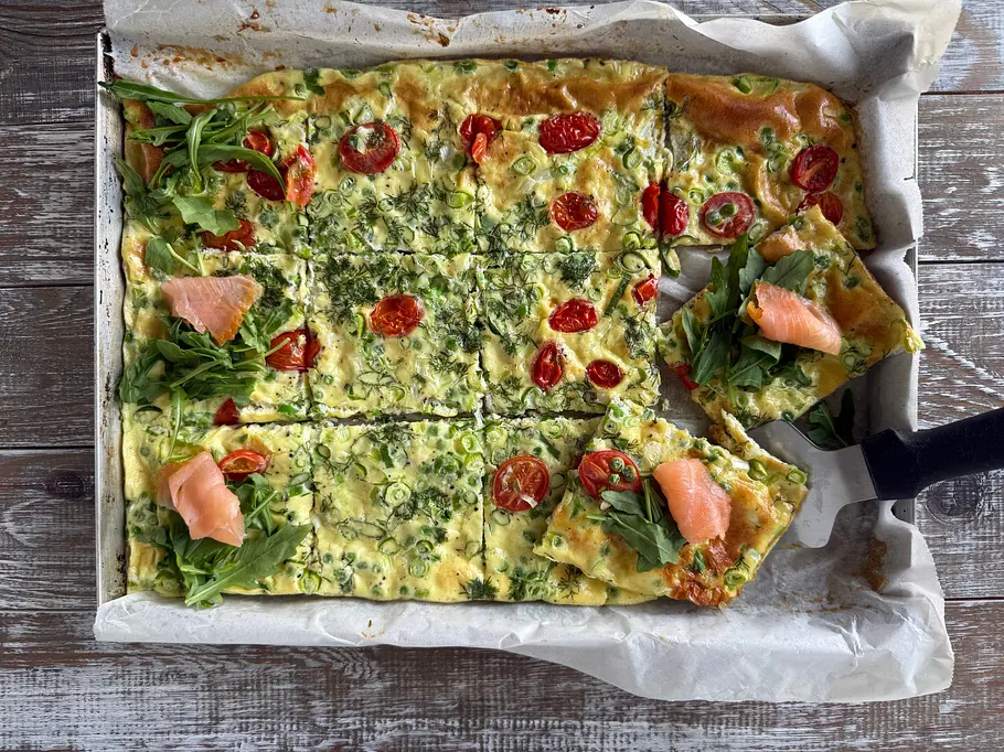 Auf einem Backblech liegt ein in Stücke geschnittenes Omelette mit Tomaten und Kräutern, teils mit Rucola und Lachs belegt.