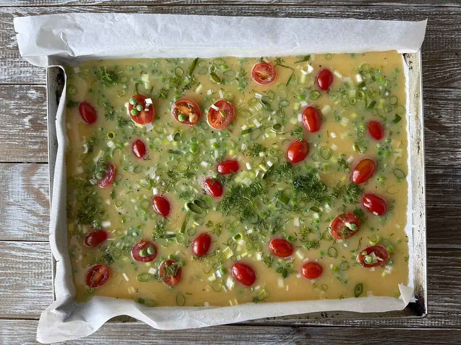 Auf einem mit Backpapier ausgelegten Blech liegt eine flüssige Eimasse mit Tomaten, Frühlingszwiebeln und Kräutern.