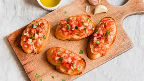 Ein Holzbrett mit vier Scheiben Bruschetta und 2 Knoblauchzehen. Dahinter ein Schälchen mit Olivenöl. - Foto: LECKER @ Bauer Media Group / mithilfe von KI generiert Ein Holzbrett mit vier Scheiben Bruschetta und 2 Knoblauchzehen. Dahinter ein Schälchen mit Olivenöl. - Foto: LECKER @ Bauer Media Group / mithilfe von KI generiert