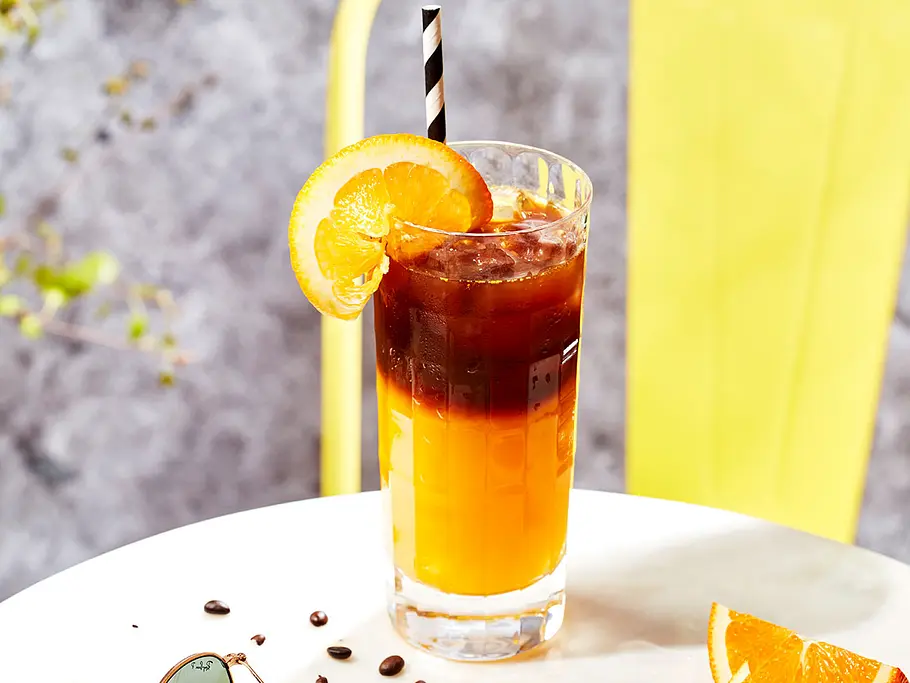 Ein Glas halb gefüllt mit Orangensaft, halb mit Kaffee und Eiswürfeln. Garniert mit einer Orangenschale und einem gestreiftem Strohhalm.