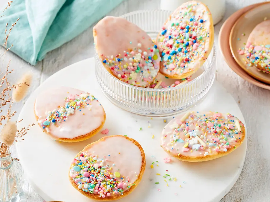 Bunte Ostereier aus hellem Mürbeteig mit zartem Zuckerguss und farbenfrohen Zuckerstreuseln, dekorativ auf einer weißen Platte und in einer Glasschale angerichtet.